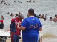 Jovem na missão Jesus no Litoral rezando com pessoas na praia.