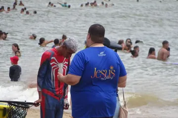 Jovem na missão Jesus no Litoral rezando com pessoas na praia.