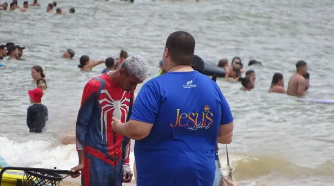 Jovem na missão Jesus no Litoral rezando com pessoas na praia. ?? 