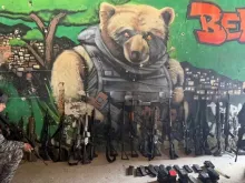 Policiais com parte da apreensão de fuzis da Operação Contenção em uma parede com grafite de um urso aramado que faz referência ao líder do Comando Vermelho (CV), Edgard Alves de Andrade, conhecido como "Doca" ou "Urso"