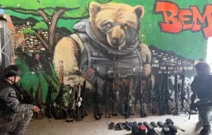 Policiais com parte da apreensão de fuzis da Operação Contenção em uma parede com grafite de um urso aramado que faz referência ao líder do Comando Vermelho (CV), Edgard Alves de Andrade, conhecido como "Doca" ou "Urso"