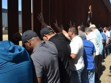 Sacerdotes scalabrinianos rezam diante do muro que separa México e Estados Unidos. Crédito: Cortesia Casa do Migrante em Tijuana.